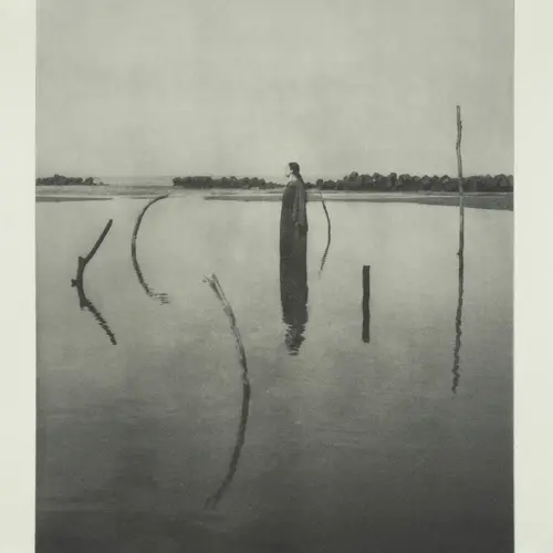 Black and white photograph of a calm, wide body of water under a hazy sky, with thin, curved sticks protruding from the water's surface. A figure stands in the centre.