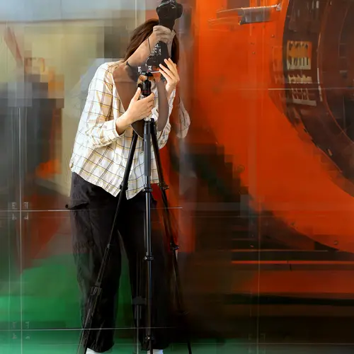 A mock up of a collapsed AI image generated from previous iterations of the Variations project. Colourful distorted images of a person stood with a camera on a tripod in front of an orange sculpture.