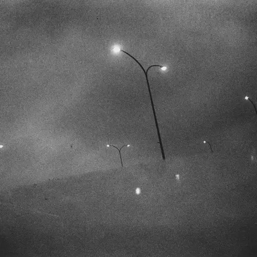 A black and white photograph of a hazy, fog-filled scene with street lamps barely visible.