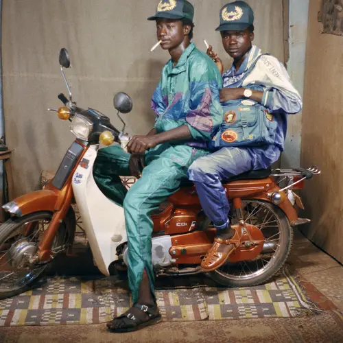 Two men sit on a motorcycle inside a studio.
