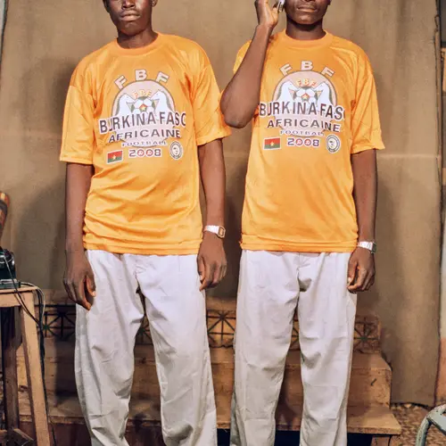 Colour photograph of two men, wearing  matching orange Burkina-Faso t-shirts and beige trousers.  One of the men wears a blue cap whilst the other has a  phone raised to his ear.