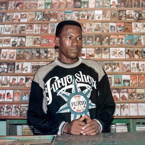 Colour photo of a man posing in front a wall full of shelved cassette tapes.