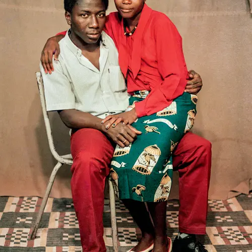 Colour photograph of a couple. The man  sits on a chair with his partner sitting on his leg
