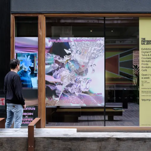 A man stands in front of a screen that shows an image where his head is changed into an alien-like creature, while a vinyl print of a lightly coloured computer avatar partially covers a large window