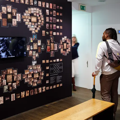 A person looks at a wall covered in photographs arranged in a pattern around a central screen