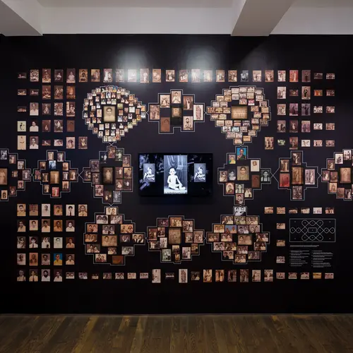 A dark wall featuring lots of printed photographs arranged in a pattern, around a central monitor