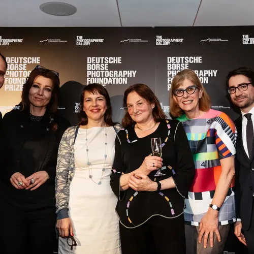 Group photograph of five people, standing in a room, watching something on the other side of the room