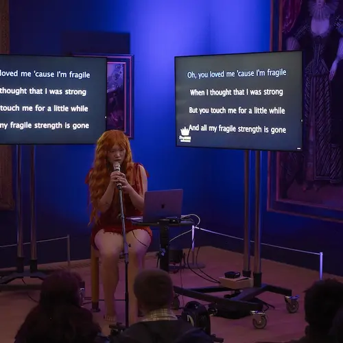 An image of the artist in a pink wig performing in front of two monitors showing song lyrics