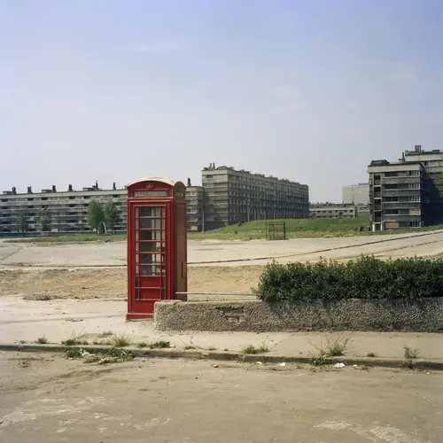 Colour photograph of a red phonebox on the street.  Blocks of flats can be seen In the distance.