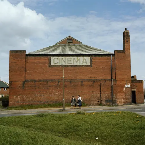  Colour photograph of a building with the word  ‘Cinema’ engraved on it. Two women can be seen standing  in front of the building.