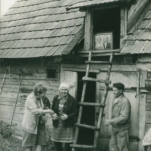 A vintage black and white photograph Zofia rydet stood outside of an old countryside cottage typical of the Polish village. An elderly couple stands with her smiling warmly.