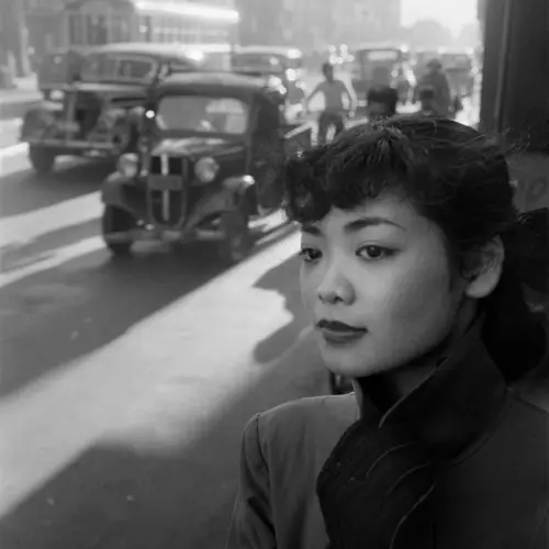 A black and white photo of a misty city street. A young woman's face is in the foreground looking out into the road.