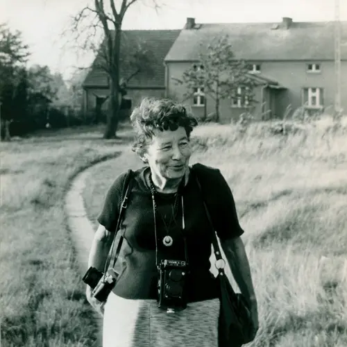 Black and white photograph of Zofia Rydet walking down a path leading from a traditional country cottage. She wears a dark blouse and has a camera on a strap slung around her shoulder.