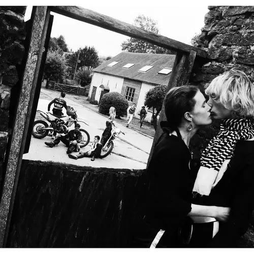 A black and white image of a couple kissing in the foreground. They are stood in a dilapidated buidling and through the window, there are several people on motorbikes riding on the road.