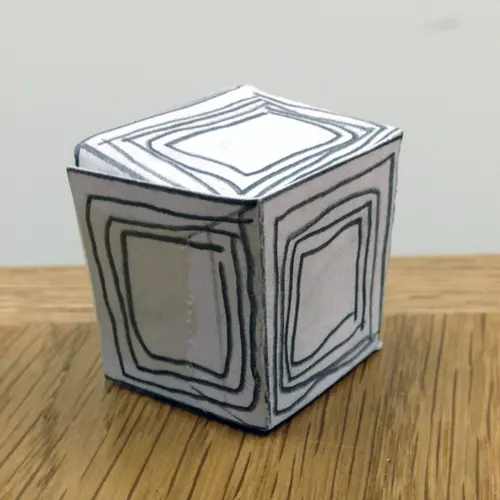 Small cube made of paper card with concentric squares drawn in pencil on each side.