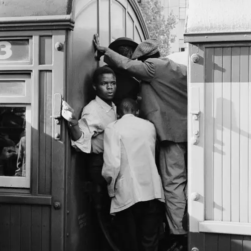 Black and white photograph of several men and boys stood between two train cars as they hold on to anything they can.