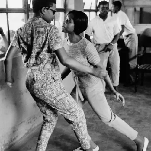 Black and white photograph a man and woman dancing together in a room. Two other men are stood in the distance. 