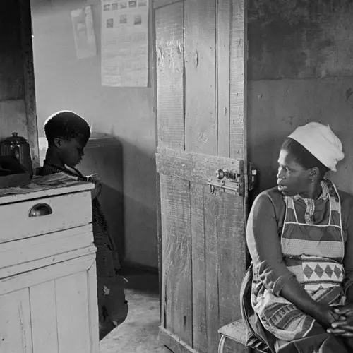 Black and white photograph of a mother sat down as she looks over to her son stood in the doorway looking sullen. 
