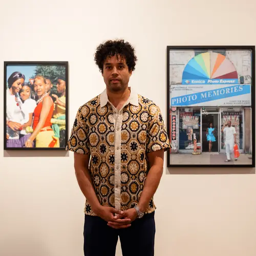 Portrait of Johny Pitts standing in a gallery space with 2 of his images on the wall behind him