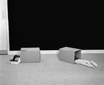 Black and white image of a girl lying on the floor with 2 boxes. Half of her torso is showing in one box and the lower half of her is showing in another box