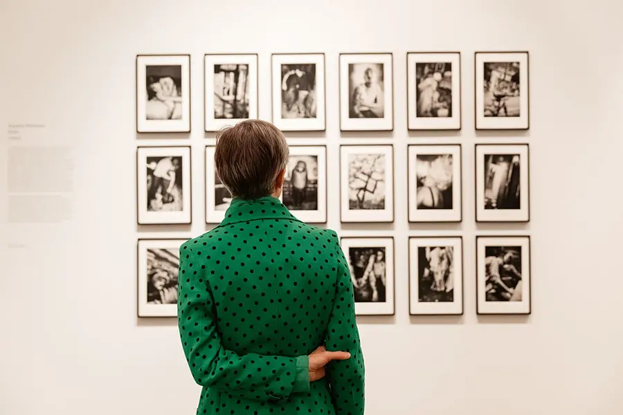 A woman wearing a green jacket stands looking away from camera at a wall of photographs