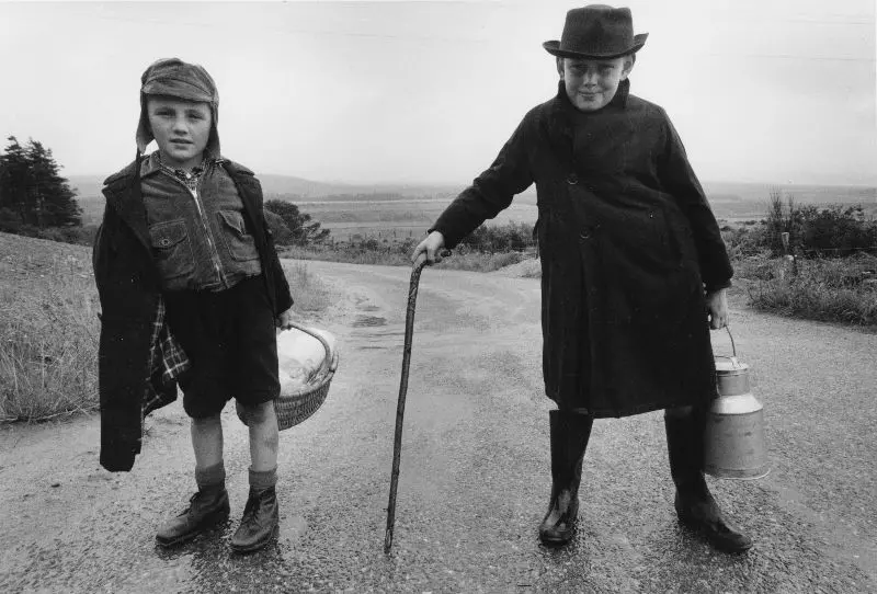 Black and white image of two boys near Inverness, Scotland. Boy on left wearing a hat, jacket and coat, shorts and boots and holding a basket. Boy on right wearing a hat, overcoat, boots holding a cane/walking stick in one hand and a metallic canister in the other 