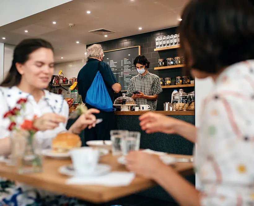 Image of The Photographers' Gallery Cafe. Two people having a conversation and enjoying lunch whilst sitting down at the table, with water, sandwich, teacup, red flowers. Centre of image has a cafe staff wearing a face mask, processing payment for a customer. Customer is wearing a short sleeve dark navy shirt, with a royal blue drawstring bag over right shoulder