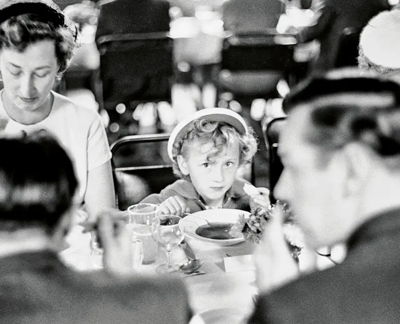 Black and white close up cropped image of someone holding a spoon in their hand with a bowl and glasses on the table 