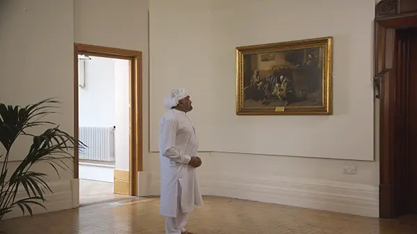 A man dressed in a white shalwar kameez and head covering stands in front of a painting in a gallery.