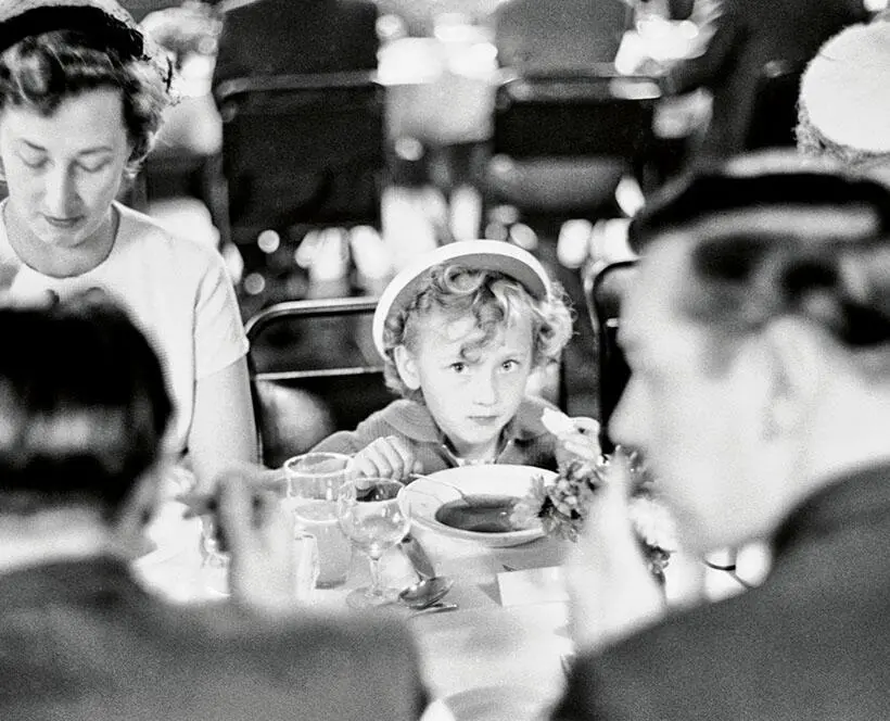 Black and white close up cropped image of someone holding a spoon in their hand with a bowl and glasses on the table 