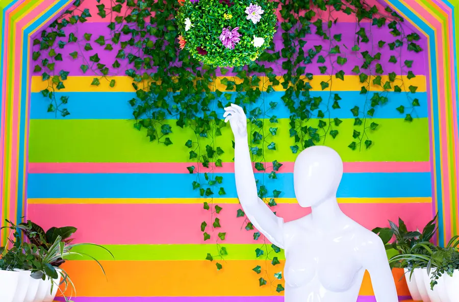 A white glossy mannequin holds its arm up in front of a colourful striped background, with potted plants