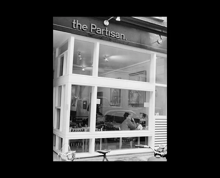 Cover of a booklet, showing a black & white photograph by Roger Mayne, showing the Partisan café window, with three men inside having coffee