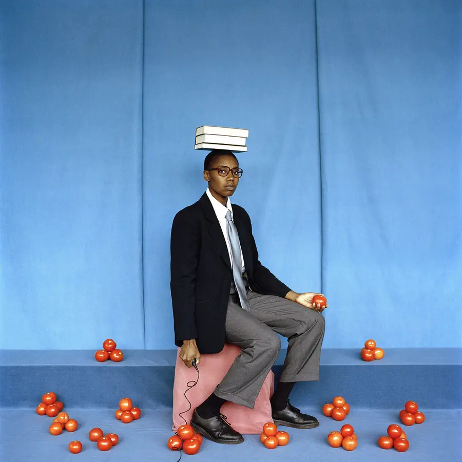 photograph of a woman dressed in a men's suit seated on a pink stool with a book on her head with tomatoes on the floor by Silvia Rosi