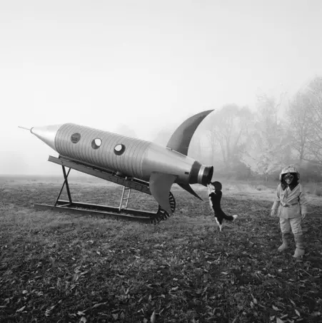 Black & white image of a child in astronaut costume standing next to a rocket maquette with its dog