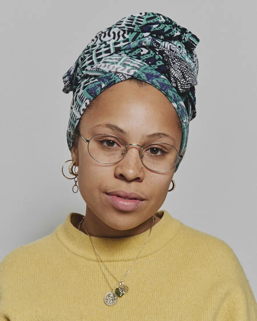 Portrait of woman wearing a colourful head wrap, yellow jumper and glasses. 
