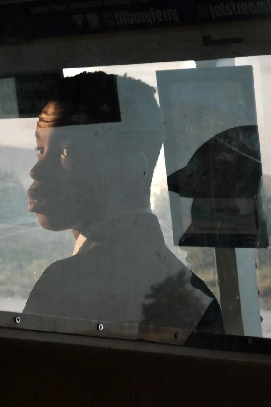 Colour photograph of a young person sat behind a glass screen, looking over their left shoulder towards the camera. There is a reflection of another person in the glass.