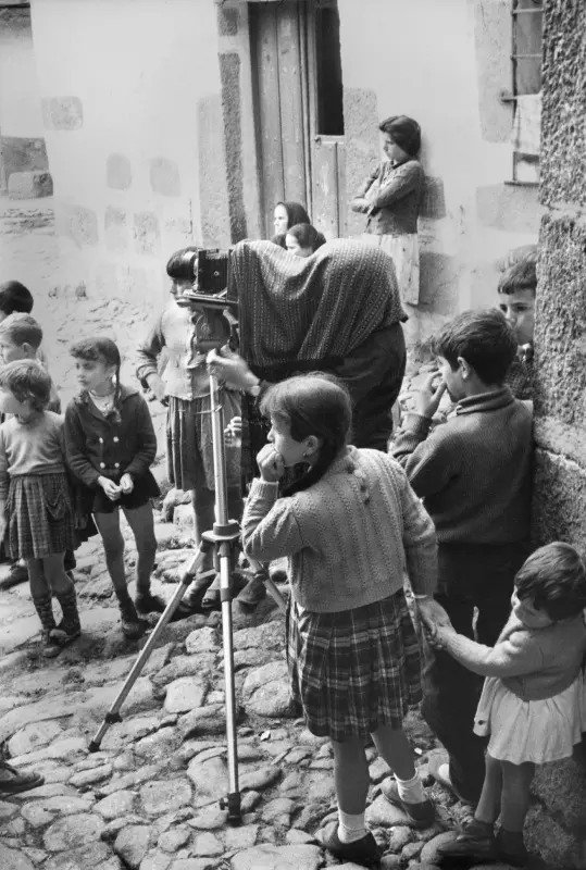 Black and white photograph of a street scene where a woman is in the centre of the image using a large format camera on a tripod, looking through it under a dark cloth. Around her are young children in the street watching.