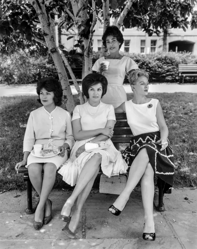 Black and white photograph of four women, three women are sat with their legs crossed and one woman is stood behind them. There is a tree in the background, immediately behind the standing woman