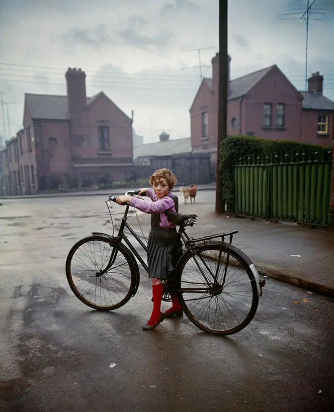 Young girl stood still holding a large bike, which looks too big for her to use. Street scene behind her. She is looking at the camera