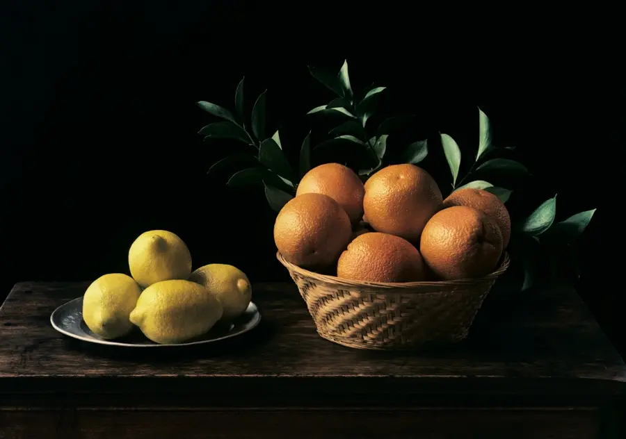 Colour photograph of a still life arrangement of a bowl of oranges on the right and a plate with some lemons on the left