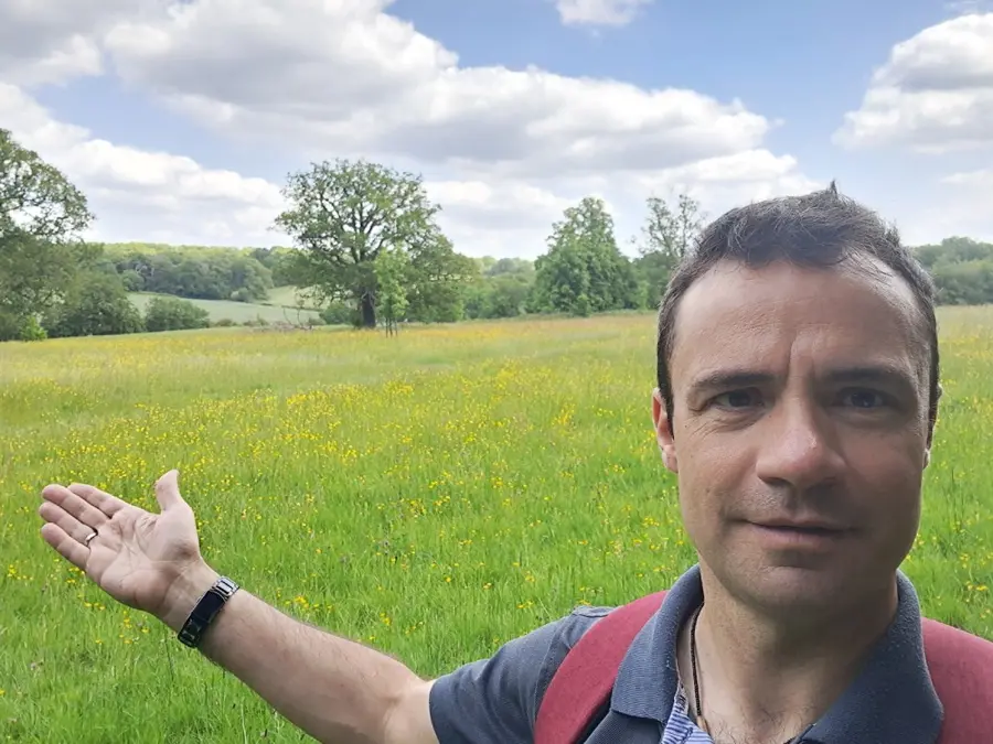 Colour photograph of a person standing infront of a green field with trees and blue sky