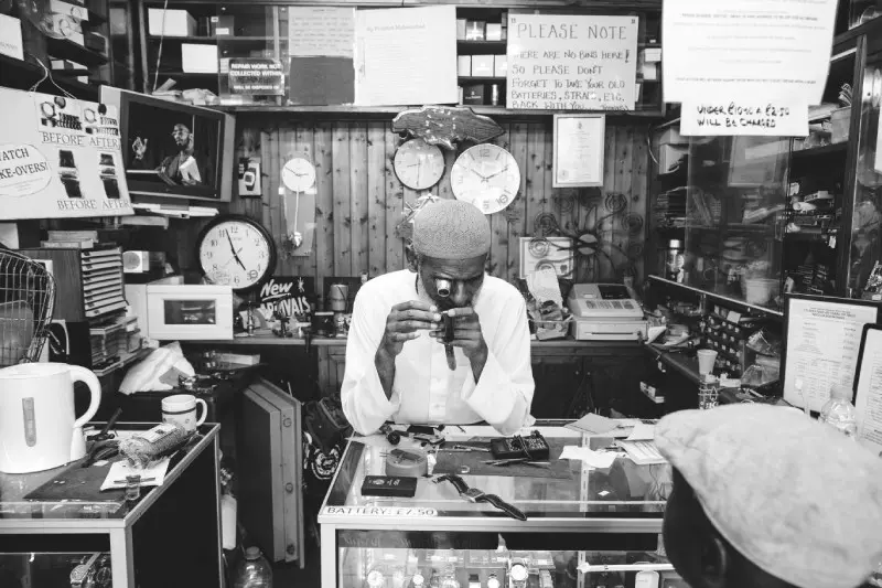 Black and white photograph of a person in a shop looking through a magnifier at a watch they are holding