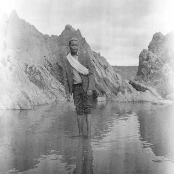 Black and white photograph of a person standing with feet submerged in water and trouser legs rolled up