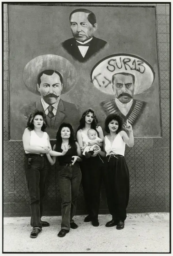 Black and white photograph of four women, one of whom is holding a baby in her arms. They are stood in front of a wall mural of three men