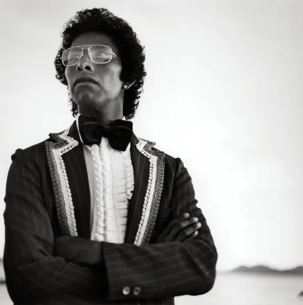 Black and white photograph of a bespectacled man in a suit and bowtie stood with his arms crossed. He looks off into the distance