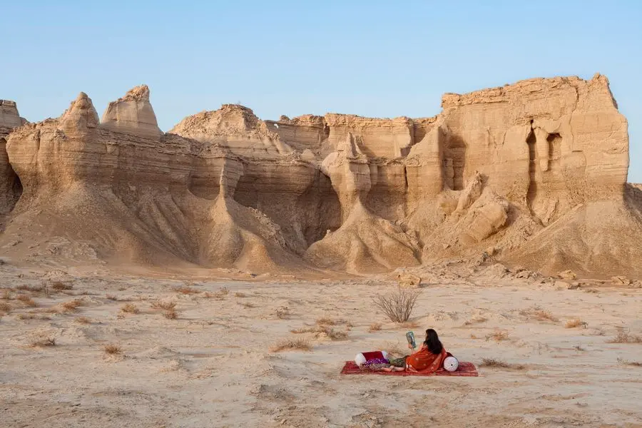 A sandy desert landscape under a blue sky . A person sits on a red rug on the floor.