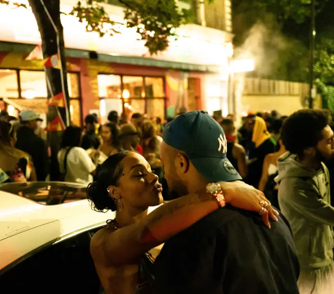 Candid photo of a couple kissing against a car with a crowd of people in the background
