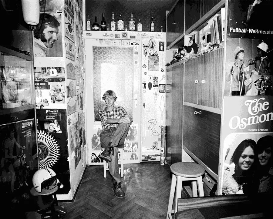 Black and white image of a young person sat inside a domestic room turned toward the camera.