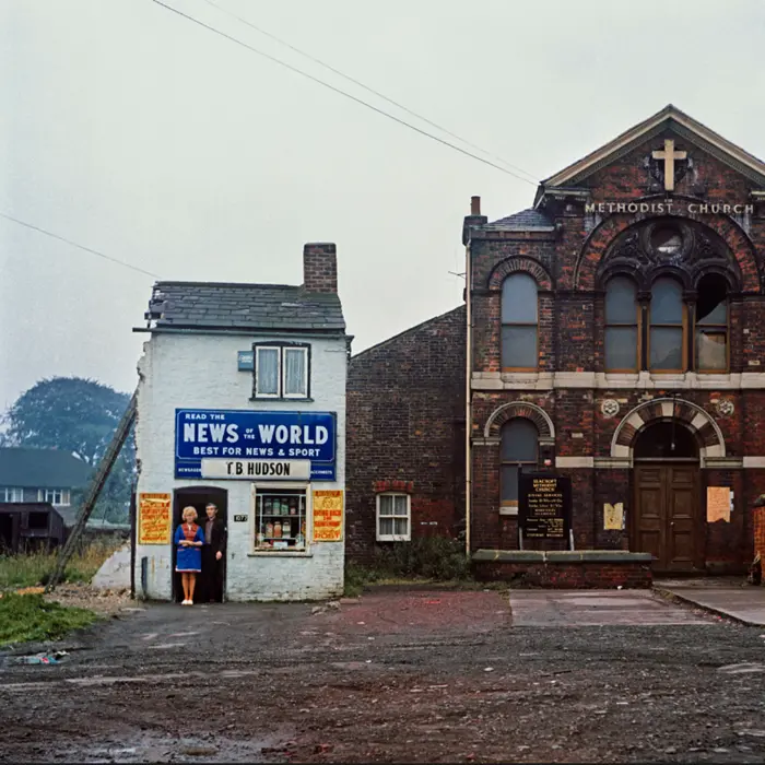 Colour photo depicting a man and woman stood in the doorway of their small shop which is next door to a brick church.