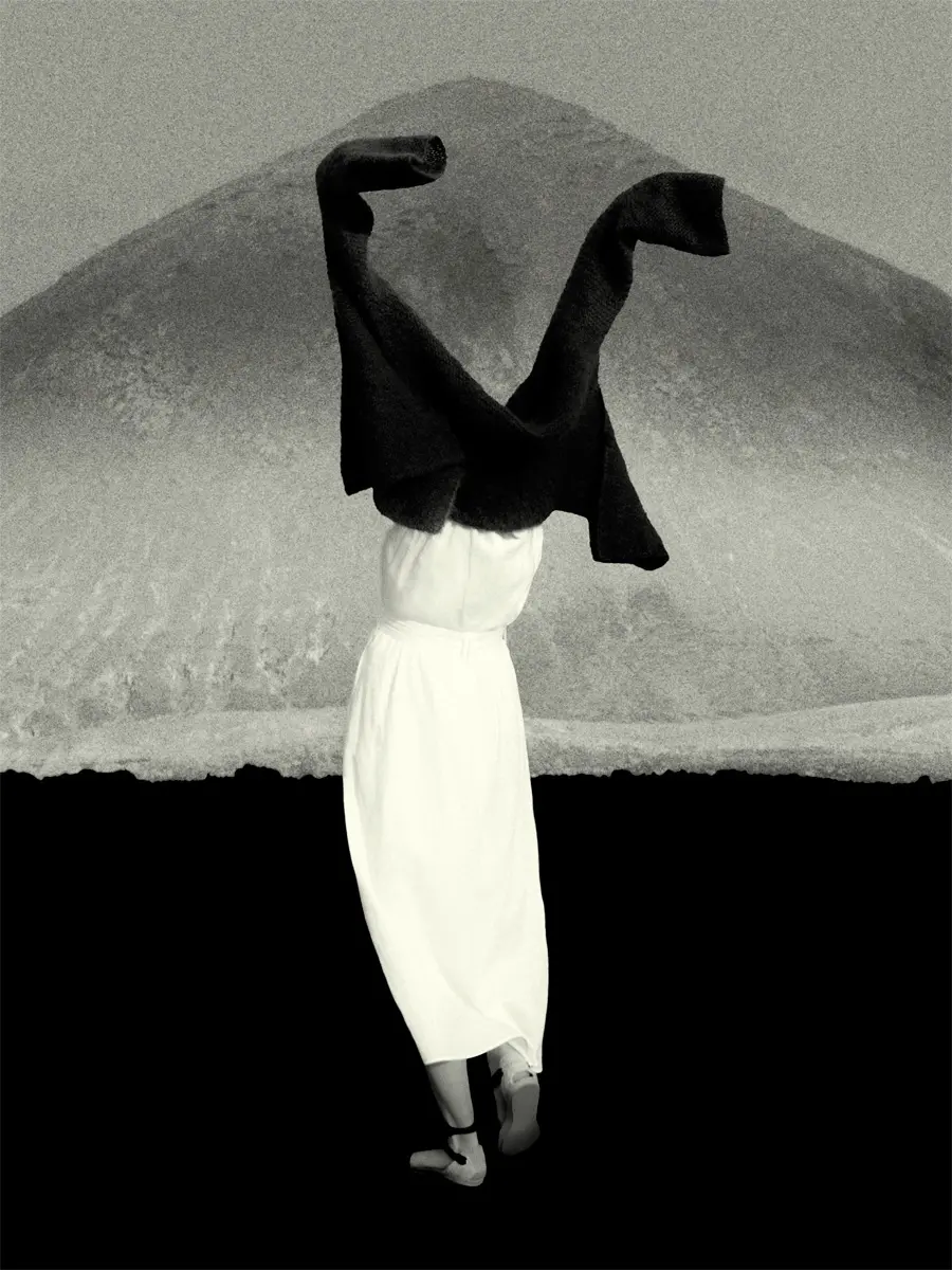 A black and white photograph of a person in a white dress reaching their arms upwards which are draped in a jacket that obscures their face. There's a mountain in the background.